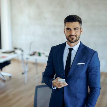 Smiling entrepreneur text messaging on smart phone in the office and looking at camera.